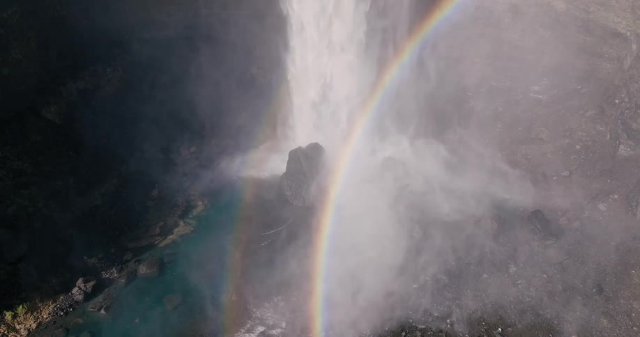 The majestic waterfall cascades down the cliff face creating a misty curtain that catches the sunlight to form a vibrant rainbow