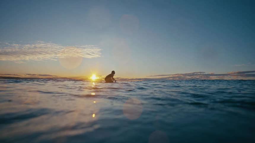A lone surfer sits on their board watching the sun dip below the horizon as gentle waves reflect the golden hues of the evening sky