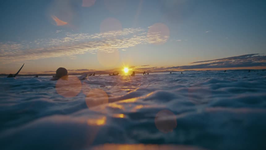 A surfer floats on the ocean as a wave passes beneath him at sunset