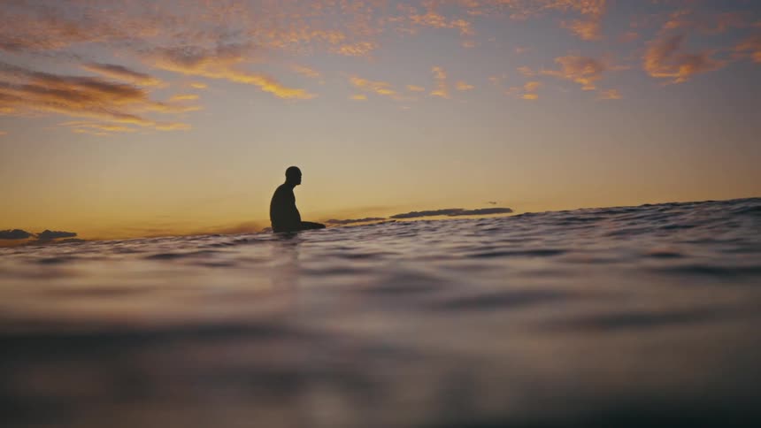 Person sitting on a surfboard in the ocean with the horizon in the background and the sky tinted orange and yellow by the sunrise