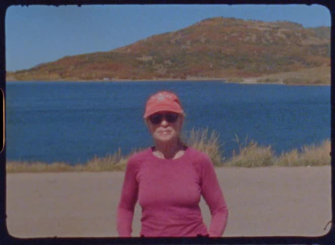 A woman wearing a hat sunglasses and a long sleeve shirt stands near a lake and waves