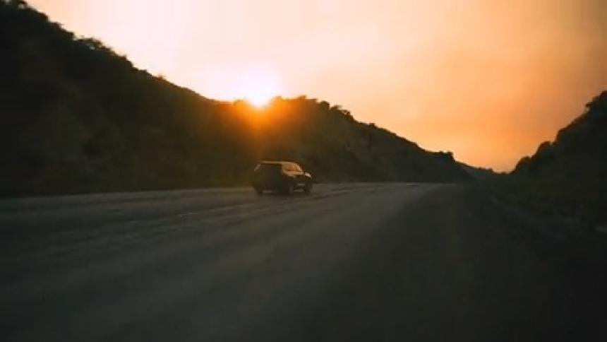 Camera races toward a black SUV driving into a smoky sunset