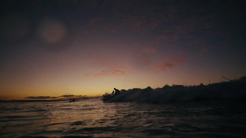 Surfers ride gentle waves at dusk silhouetted against a warm fading sunset sky