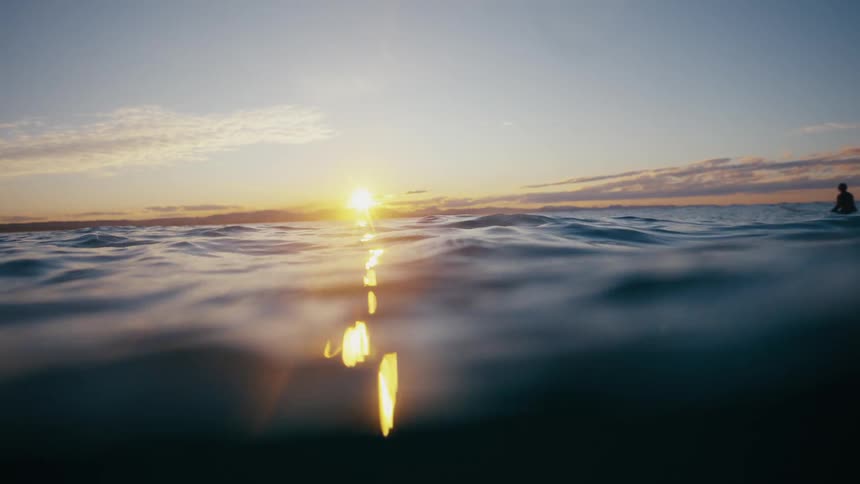 A man is floating on his surfboard in the ocean at sunrise