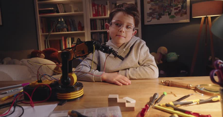 A boy sits at a desk with a robotic arm and various tools and wires