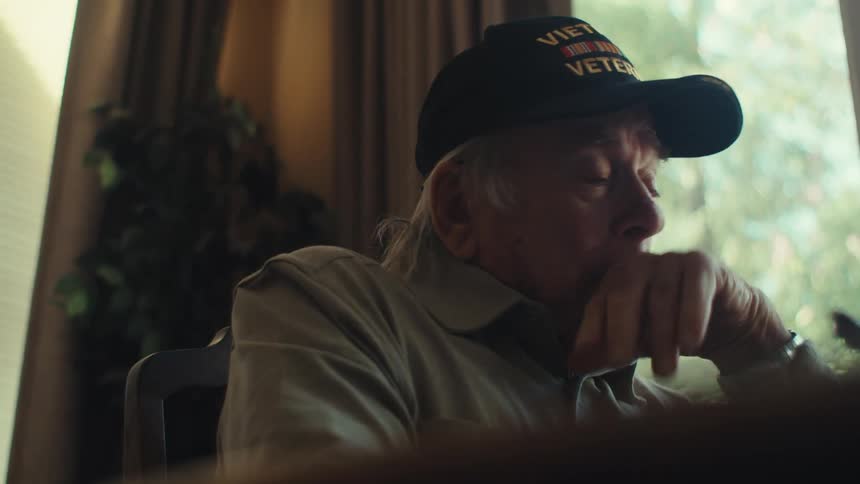 An elderly man wipes tears from his eyes as he writes a letter at home
