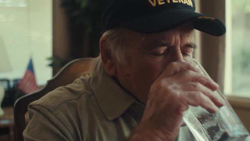 The elderly veteran wearing a blue cap and a gray shirt sips water while jotting notes in his notebook at the table