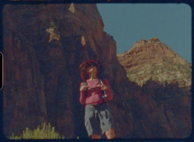 A woman in a sun hat and backpack stands at the base of a steep mountain range looking up