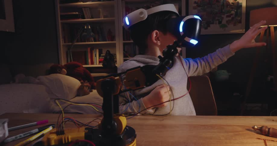 A boy wearing vr goggles operates a small robotic arm on a desk
