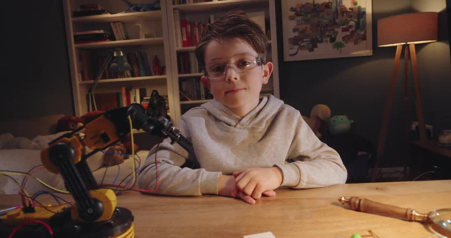 A boy wearing clear goggles sits on a desk next to a small robotic arm