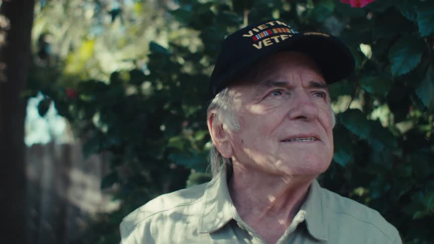 An old man wearing a vietnam veteran hat stands outdoors and sighs