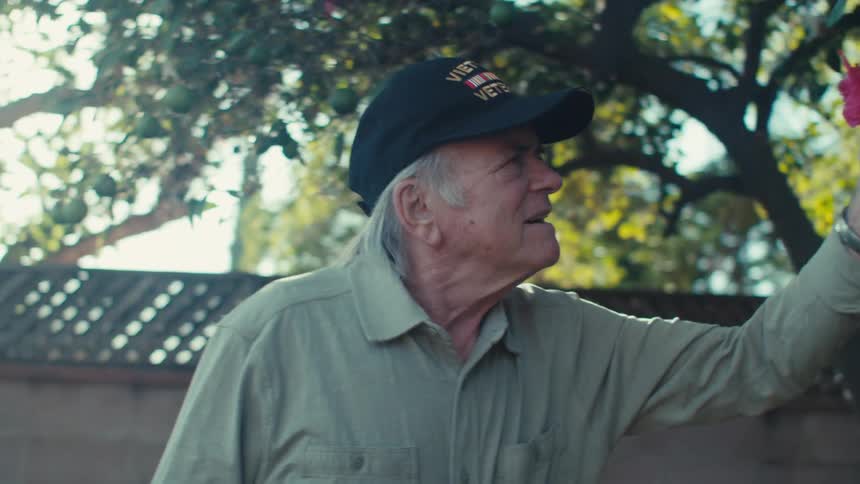 Elderly vietnam veteran holds a small american flag while smelling a flower in the garden