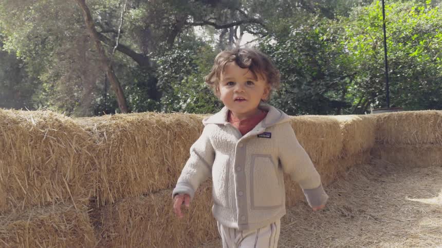 A toddler wearing a coat and sandals walks through a maze made out of hay bales