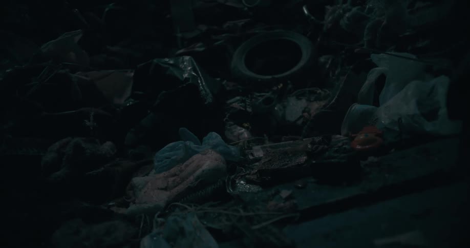 A dimly lit close up view shows a pile of garbage including a tire plastic bags and other debris