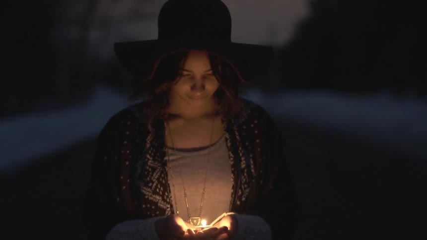 A woman wearing a large hat watching a tealight burn in her hand before looking up