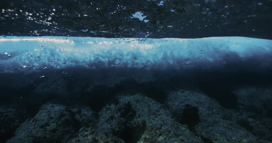 An underwater view of a wave cresting and breaking with a rocky seabed visible below