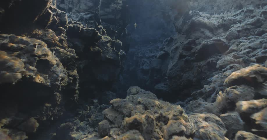 A diver swims forward through a narrow rocky chasm shafts of light illuminating the rough walls and debrisfilled bottom