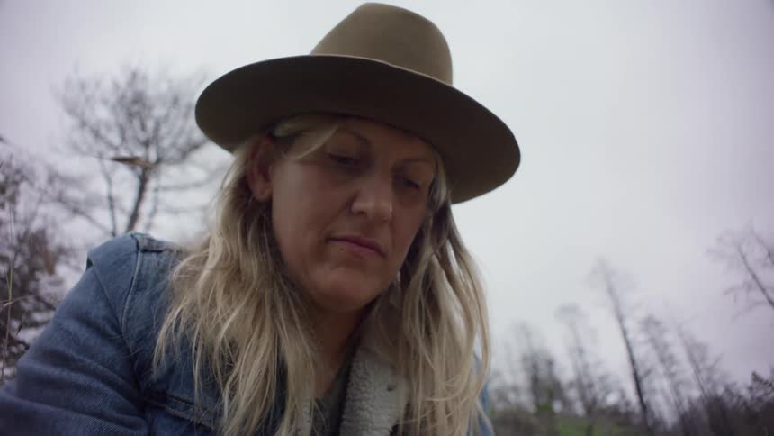 Closeup of a woman with long blonde hair wearing a hat and a jean jacket working on the ground on a hill