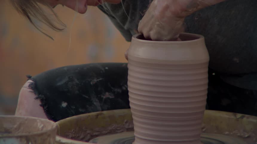 A woman works on a pottery wheel outdoors in front of a weathered wall