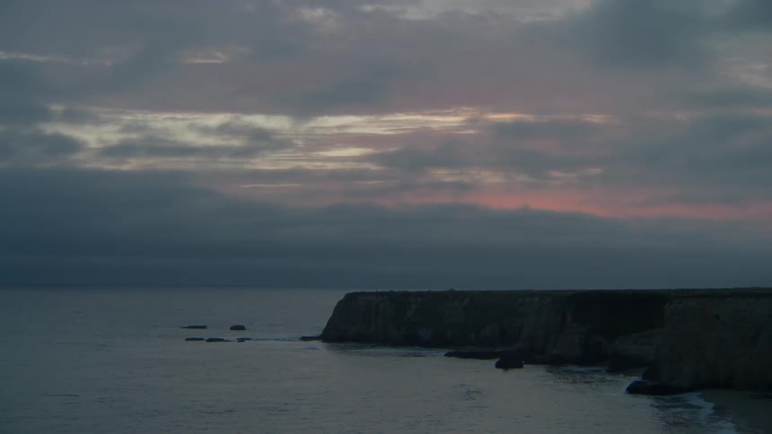Waves flow onto a beach and crash against nearby cliffs under a cloudy red tinted sky