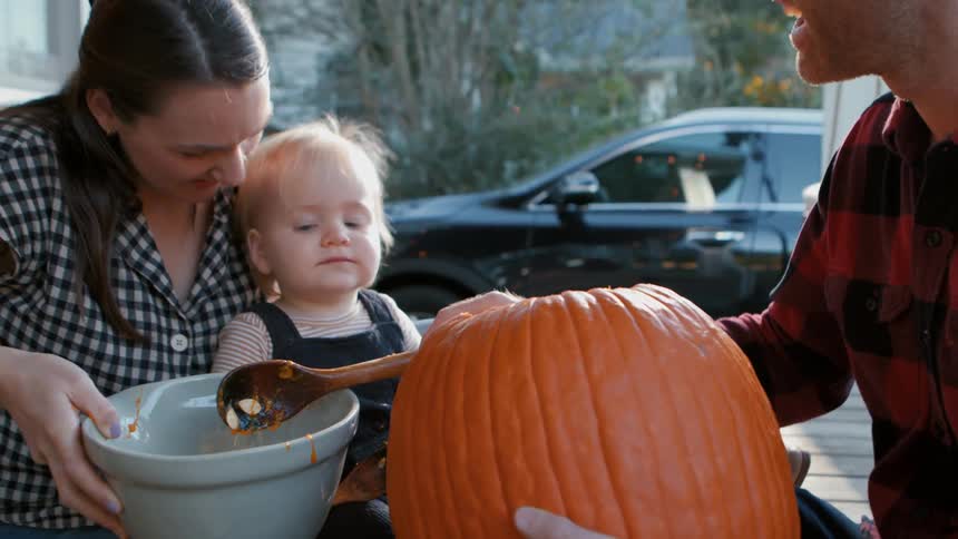 Little boy on his mothers lap looking at his father who is taking the filling out of a large pumpkin and putting it in a bowl
