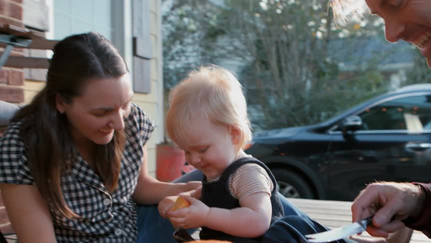 Family in backyard with father handing a piece of a halloween pumpkin to his toddler