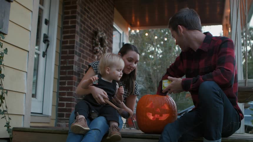 Mother sitting on stairs with young son and father next to her lighting a homemade halloween pumpkin with a match