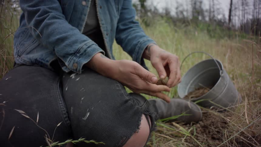 Woman sitting in a field near a filled metal bucket squeezes clay