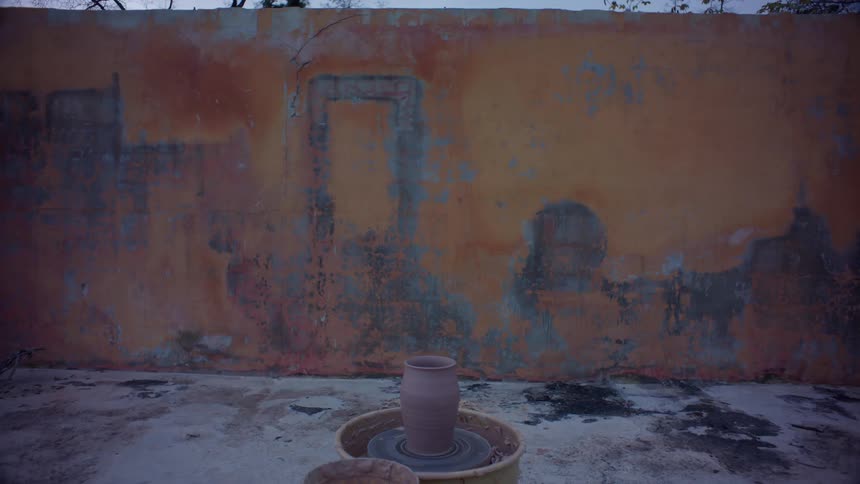 A clay vase rests on a turning wheel in front of a worn weathered wall