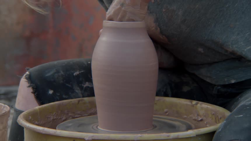 A woman works on a pottery wheel outdoors in front of a weathered wall