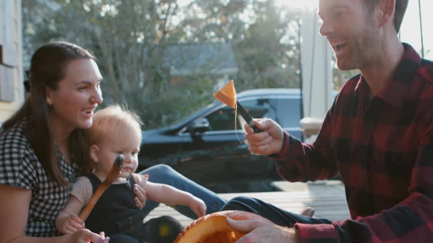 Father carving a pumpkin outdoors as mom and baby watch laughing