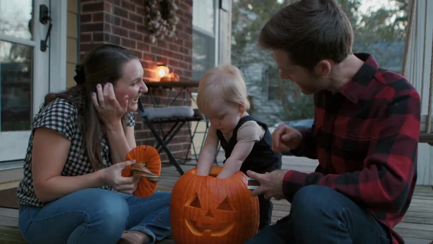 A father and mother help their child work on a pumpkin on a porch