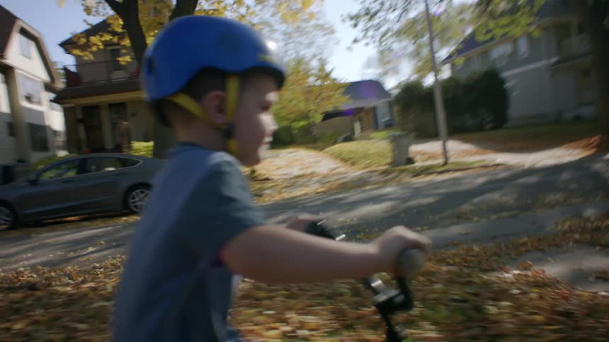 A young boy wearing a helmet rides his bike down a sidewalk covered in fall leaves