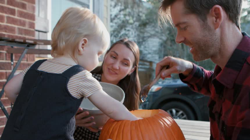 A mother and father scoop out a pumpkin as their toddler looks on