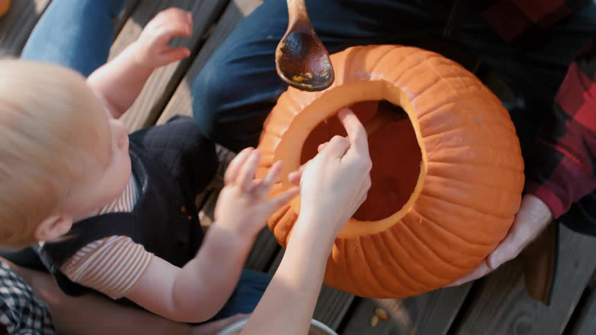 Mother and father helping baby scoop seeds out of pumpkin outdoors on a wood deck