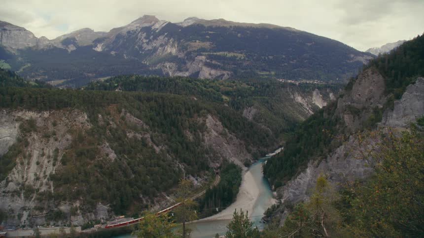 Red train passing by a riverbank in a mountain valley