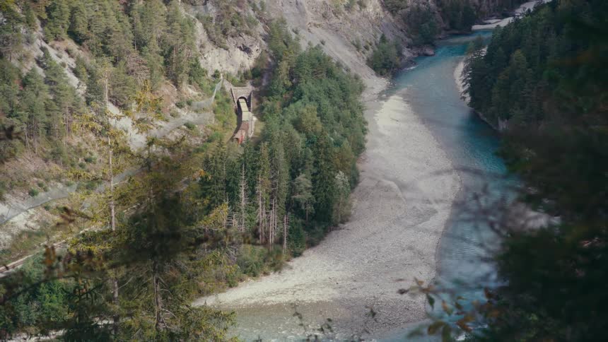 Aerial view of a train entering a tunnel beside a winding river in a narrow valley