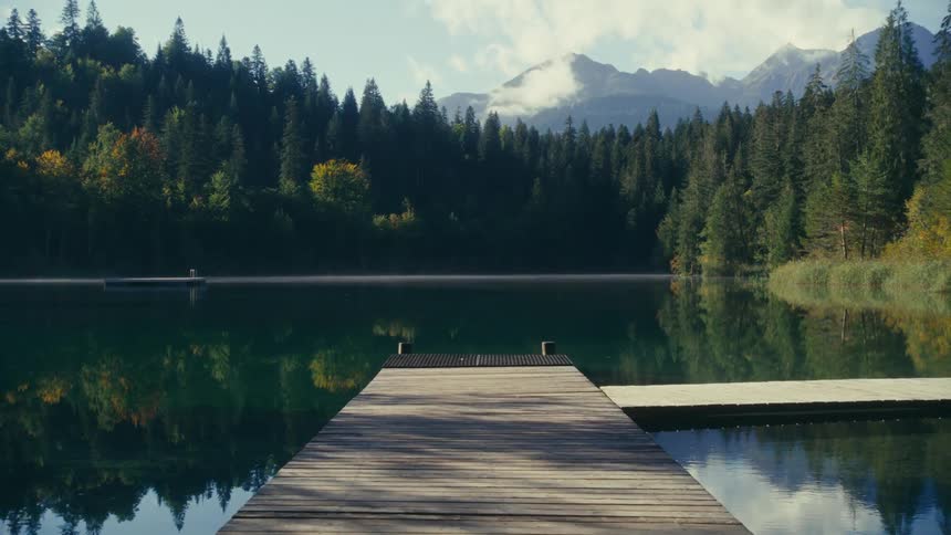 Landscape of a wooden pier on a calm lake surrounded by conifers and mountains in the background