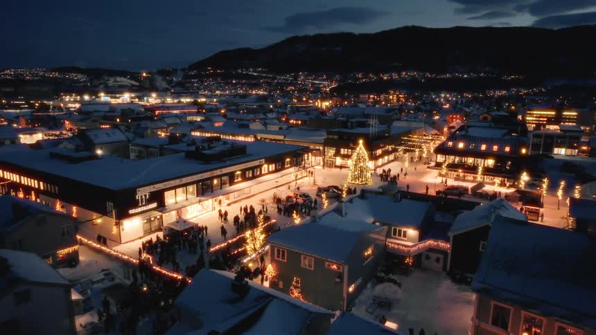 An aerial view shows a snowcovered town square decorated with christmas lights and a central christmas tree with people gathered around it at dusk
