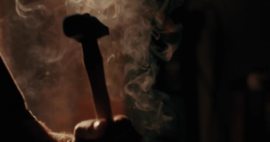 A blacksmith sets the hot head of a hammer on its handle on an anvil in a dimly lit workshop