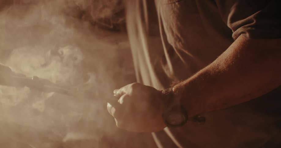 A blacksmith handles a hot smoking piece of metal with tongs in a dimly lit workshop