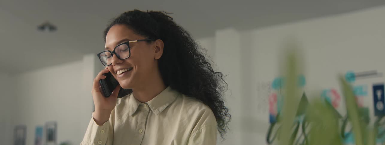 A happy woman with curly hair gestures while talking on a call in the office