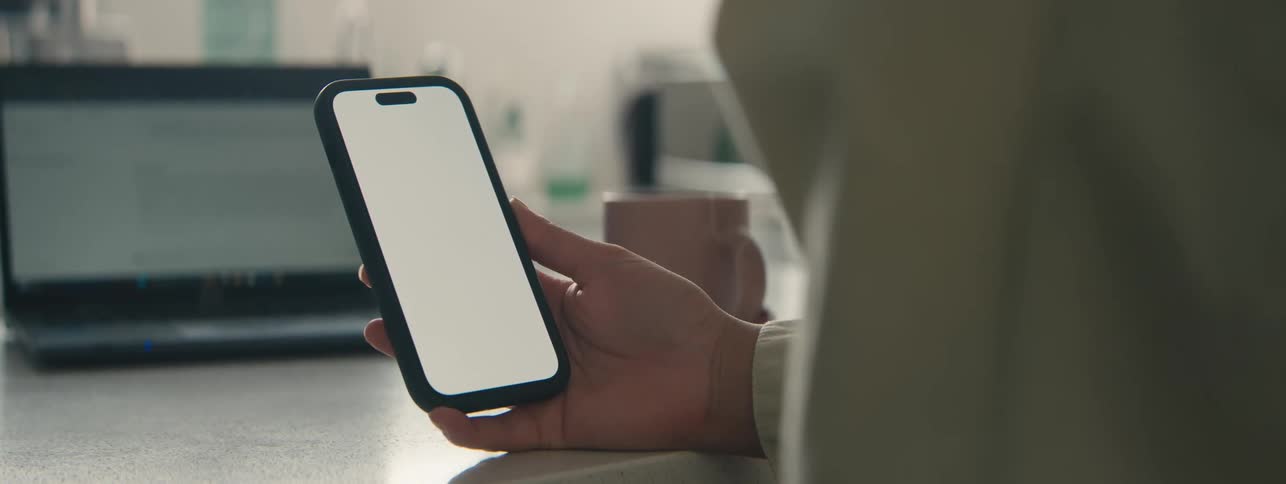 A person holds a smartphone with a blank screen with a laptop in the background