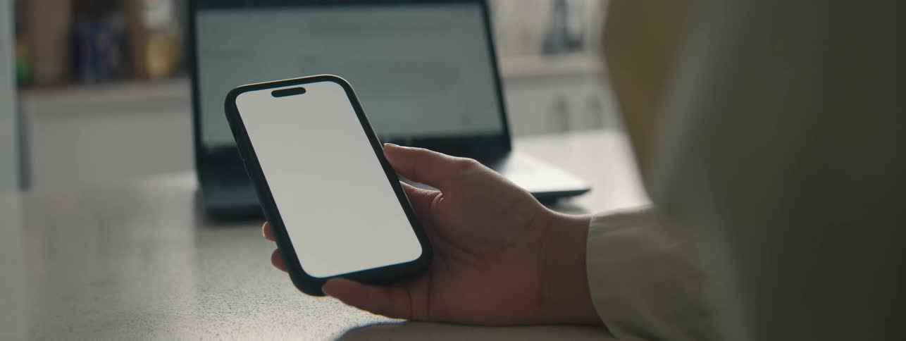 Hand holding a smartphone and in the background an open laptop on the table