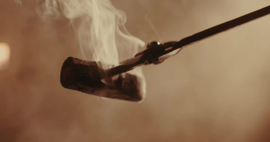 A smoking hot piece of metal is held by tongs in a dimly lit workshop