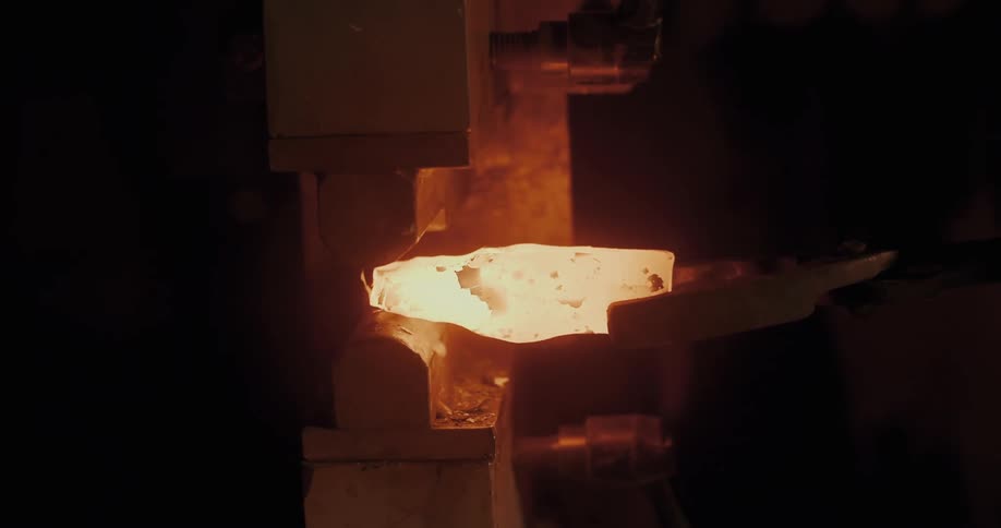A piece of redhot metal is shaped into a hammer head under a press in a dimly lit workshop