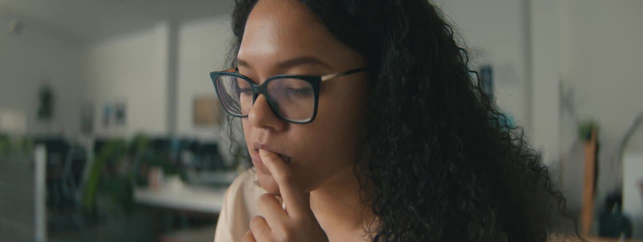 Closeup of the face of a young woman with curly hair and glasses focused on a task