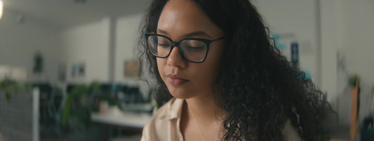 A woman with curly hair and glasses thoughtfully looks down while working
