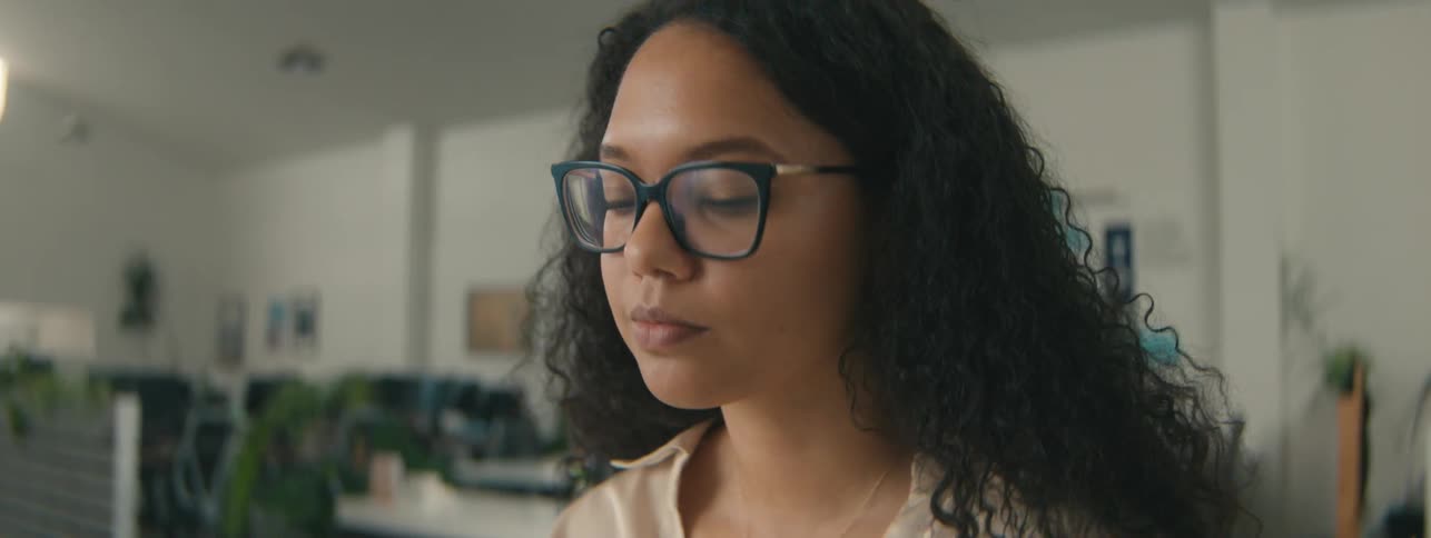 Closeup of the face of a young woman with curly hair and glasses focused on a task