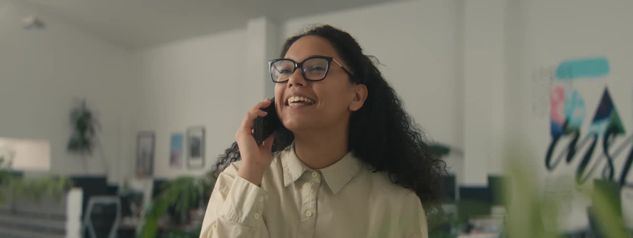 A happy woman with curly hair smiles while talking on a call in the office